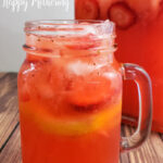 Pint sized mason jar with handle filled with strawberry lemonade with pitcher in background on wood table