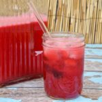 Pint of cherry limeade next to the full pitcher on a worn wood table and bamboo background