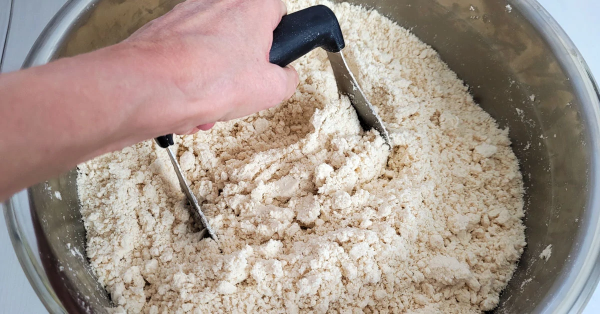 Flour and oil being mixed with a dough blender in a large metal mixing bowl.