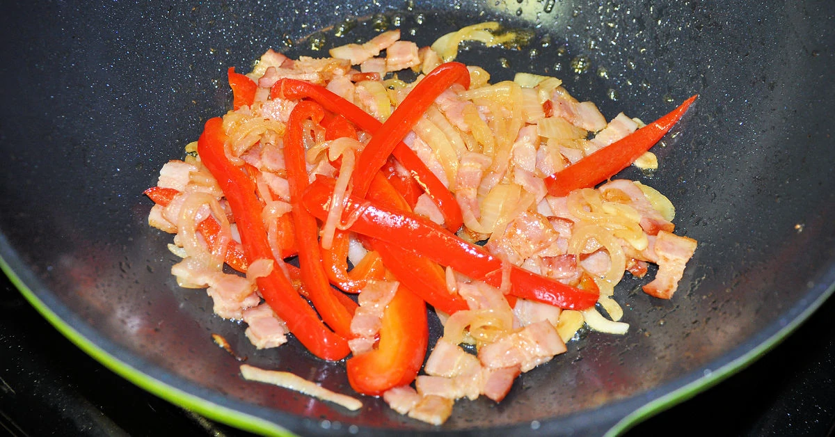 Bacon, bell pepper and onion being sauteed in a large skillet.