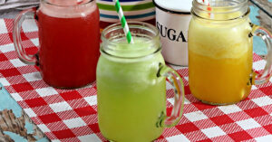 Three pint sized mason jars with handles filled with 3 types of agua fresca (watermelon, cucumber and orange) on a red and white checked table cloth.