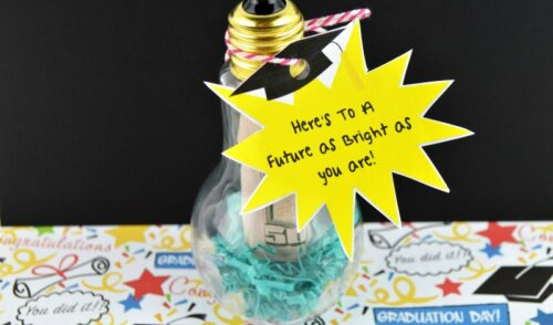Black background and graduation paper on a table with craft light bulb stuffed with blue shredded paper and a $50 with a yellow label reading, "Here's to a future as bright as you are"