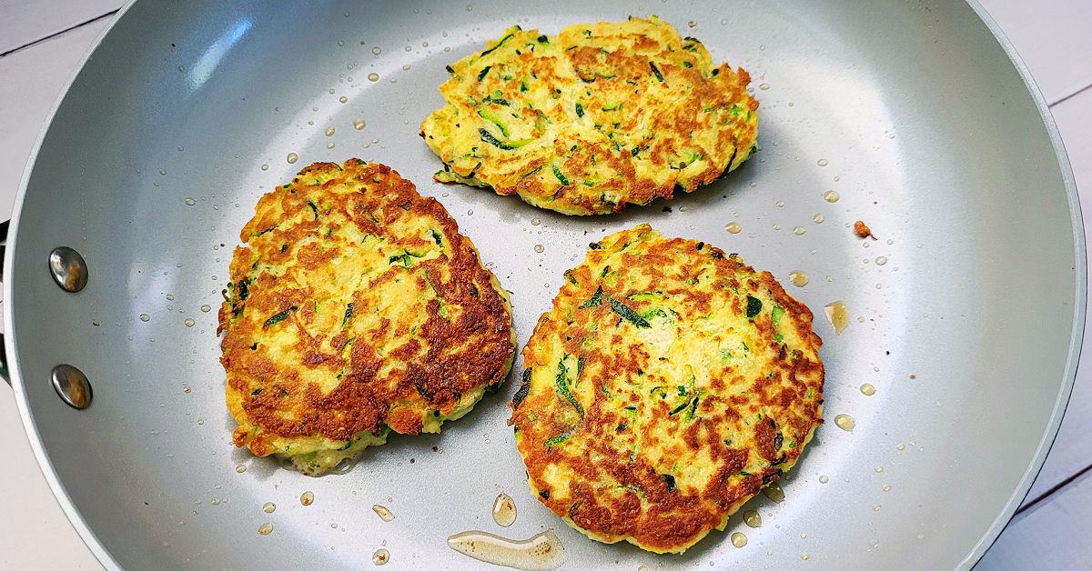 3 golden brown zucchini fritters being cooked in a ceramic coated frying pan.