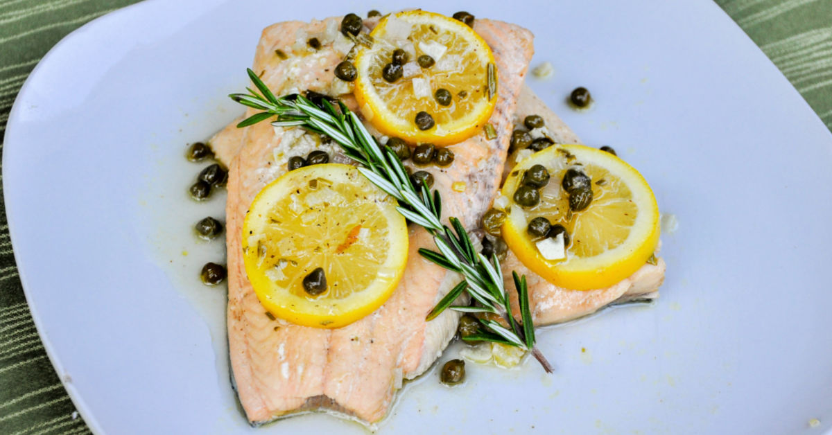 Two perfectly poached salmon filets on a dinner plate, garnished with lemon, capers and rosemary.