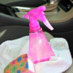 Pink plastic spray bottle of homemade upholstery cleaner on white towel next to sponge on passenger side car seat