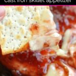 Hand dipping cracker into baked brie with pepper jam in cast iron skillet