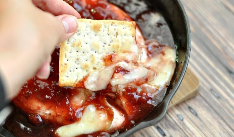 Sweet and spicy baked brie in a cast iron skillet being dipped into with a wheat cracker