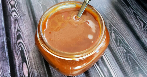 Salted caramel sauce being poured from the pan into a squat glass mason jar on a dark wood table.