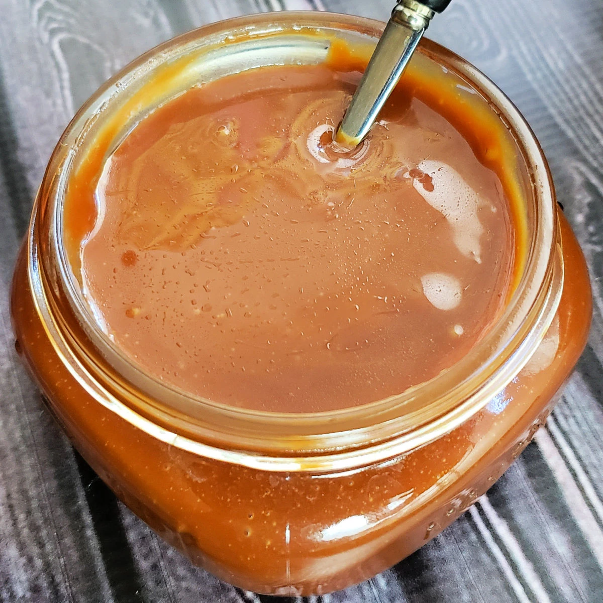 Close up shot of salted caramel sauce in a squat glass mason jar with a spoon dipped in it. Caramels are in the background.