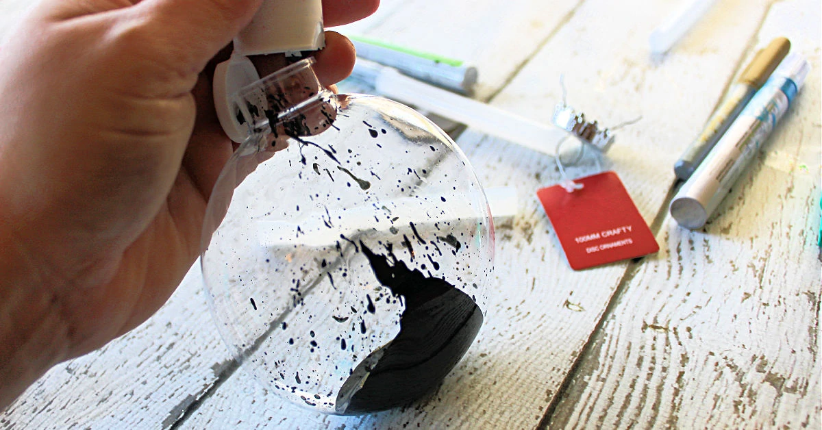 Black paint being squeezed into a clear, flat, round Christmas ornament.