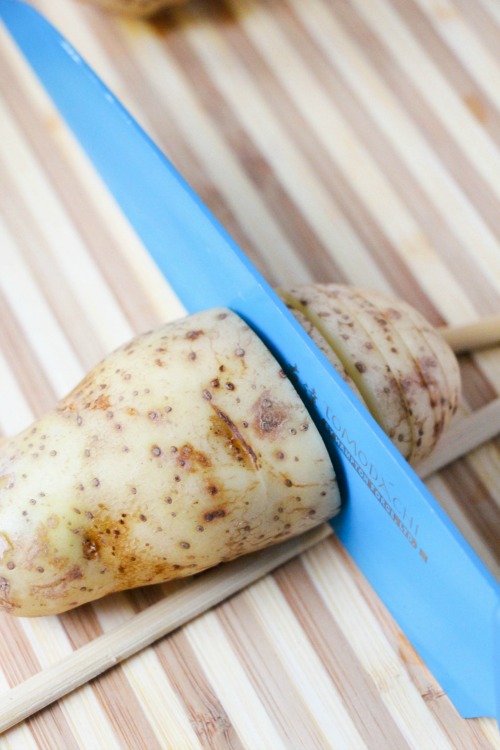 Blue knife cutting through a potato resting on chopsticks to prepare for Hasselback potatoes