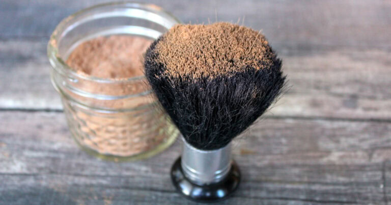 Close up of DIY bronzer on large makeup brush with jar of more bronzer in background on wood table.