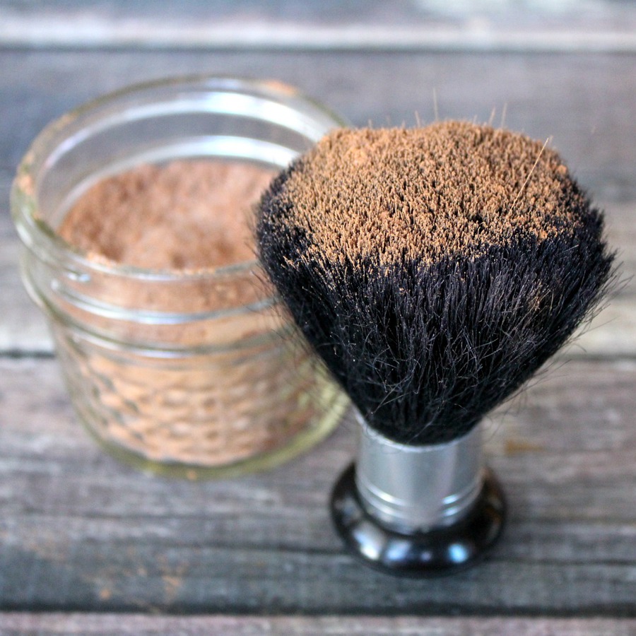 Close up of large makeup brush dipped in homemade bronzer in a glass jelly jar on a wood table