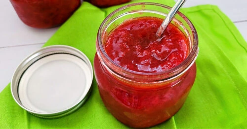 Open jar of homemade strawberry jam with a spoon in it, sitting on a green napkin.