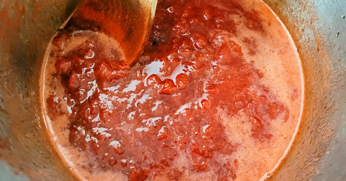 Strawberry jam being cooked down in a large stockpot and stirred with a heavy wood spoon.