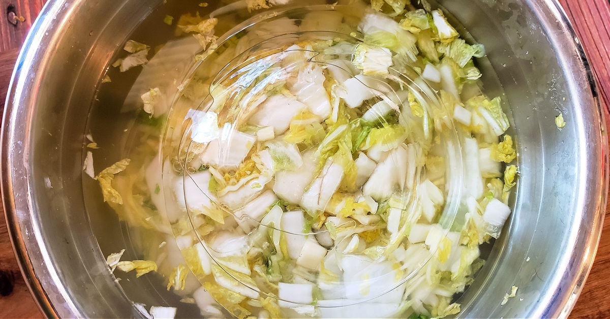 Massaged Napa cabbage covered in water in a metal mixing bowl, then weighted down with plates.