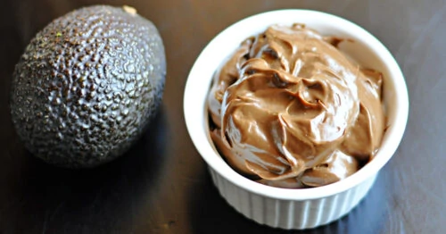 Chocolate avocado mousse in bowl with whole avocado on table.