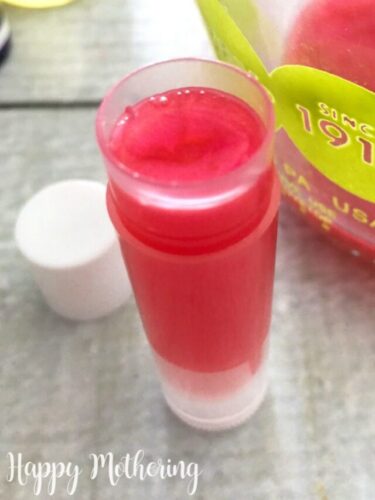 Single chapstick tube filled with melted pink tinted lip balm on a white wood table with the cap and a measuring cup of more lip balm mixture next to it