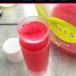 Single chapstick tube filled with melted pink tinted lip balm on a white wood table with the cap and a measuring cup of more lip balm mixture next to it