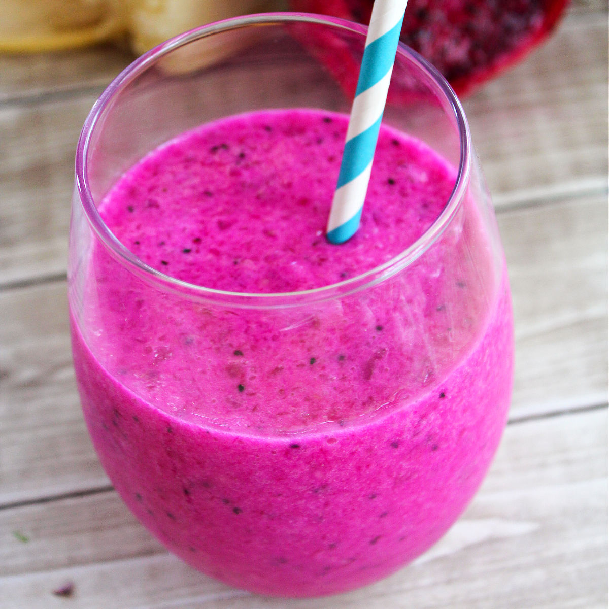 Dragonfruit smoothie in a glass jar with a white and blue paper straw on a light brown wood table background with some dragon fruit and banana behind it