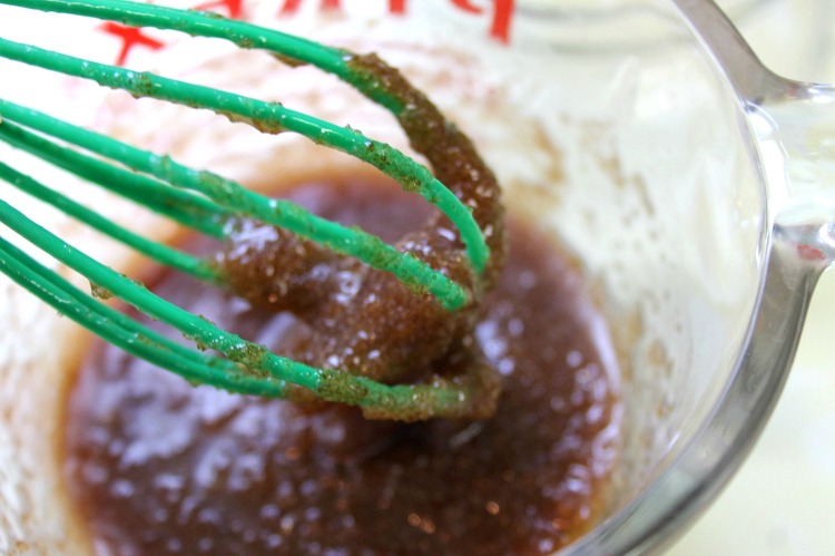 Green whisk mixing honey into brown sugar and coconut oil mixture in glass measuring cup