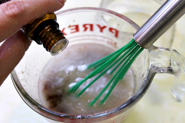 Essential oils being added to honey, sugar and coconut oil mixture in a glass measuring cup with a green whisk sitting in it