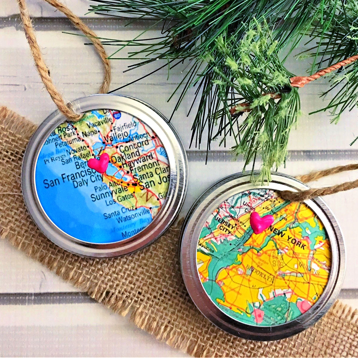 Two Mason Jar Ring Map ornaments with burlap and artificial Christmas tree branches on a vintage white wood table.