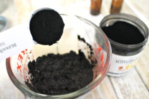Activated charcoal powder being added to a glass measuring cup containing coffee grounds, coconut oil and essential oils.