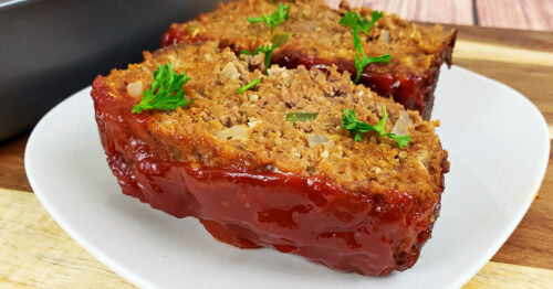 Two slices of gluten free meatloaf on a square white plate, topped with chopped parsely.