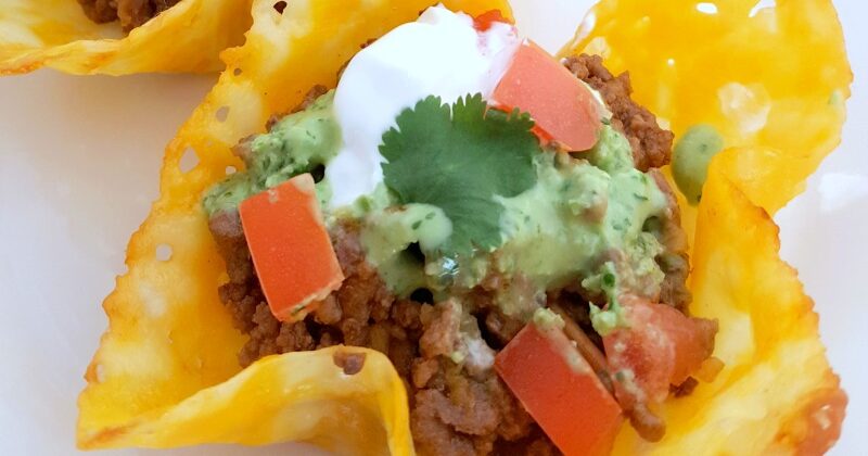 Close up of a keto taco shell on a white plate topped with ground beef, tomatoes, guacamole sour cream and cilantro