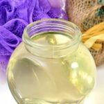 Glass jar of apple cider vinegar and essential oil hair rinse on white counter with purple sponge and decorative plant
