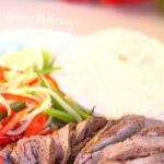 White plate of steak fajita meat, with bell peppers and onions and a bowl of sour cream with red and green bell peppers in the background