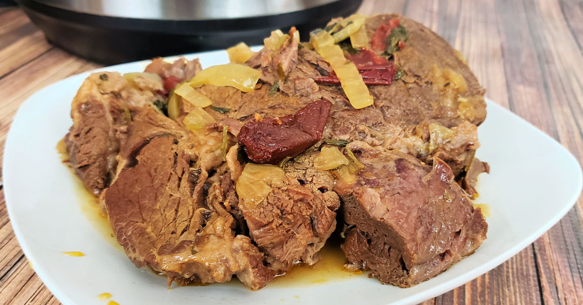 Barbacoa beef roast on white plate in front of Instant Pot after pressure cooking.