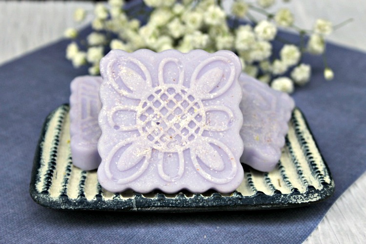 Lavender salt scrub bars on a blue and white soap dish on a blue placemat with white flowers