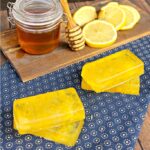 4 homemade honey lemon soap bars on a blue cloth with a white pattern on it. Behind the soap bars are a jar of honey and sliced lemon on a cutting board.
