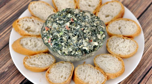 Spinach dip in a glass bowl in the center of a white plate surrounded by bread slices on a wood table