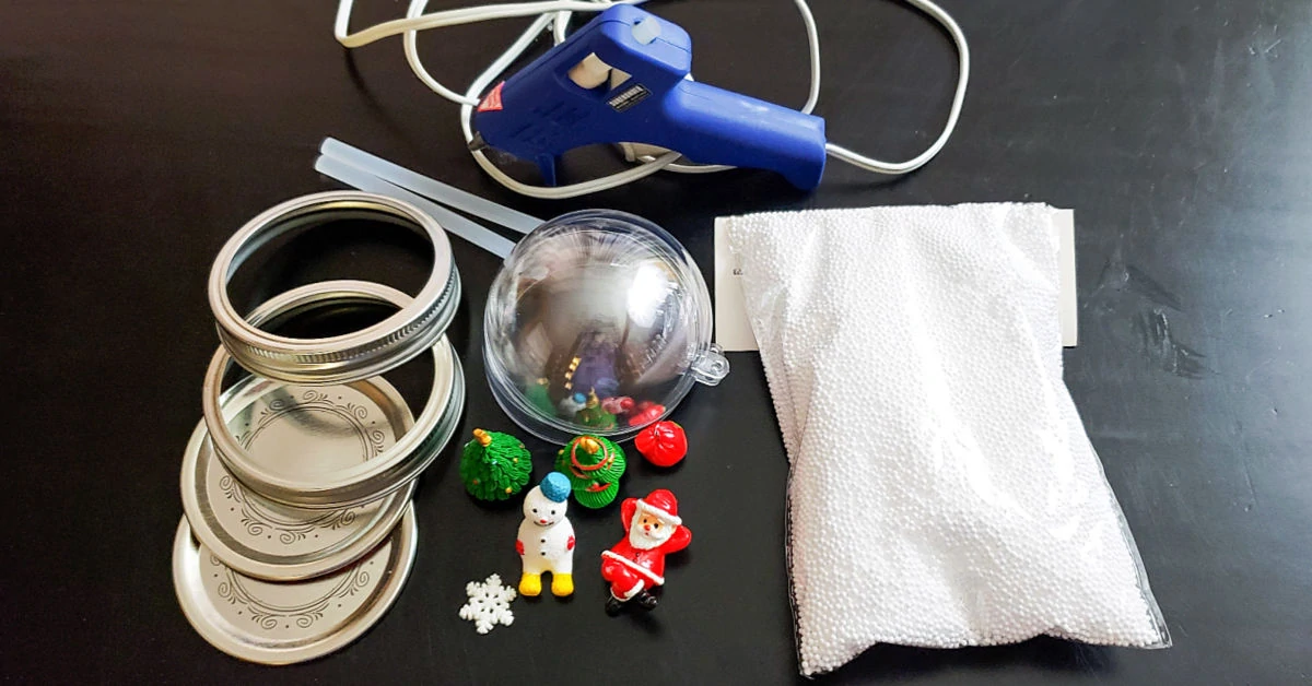 Supplies to make a snow globe mason jar topper, including mason jar lids and rings, Christmas figurines, fake snow and a hot glue gun.