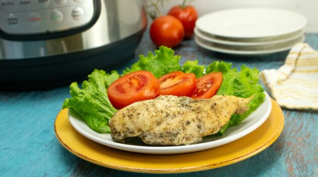 Chicken breast, lettuce and tomato on a white plate on a blue wood counter in front of an Instant Pot