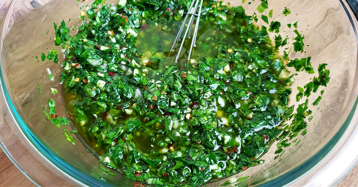 Olive oil and red wine vinegar being mixed with fresh herbs in a mixing bowl.