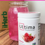 Raspberry Ultima Replenisher Container in front of quart sized mason jar full of electrolyte water in front of an iHerb shipping box on a wood table.