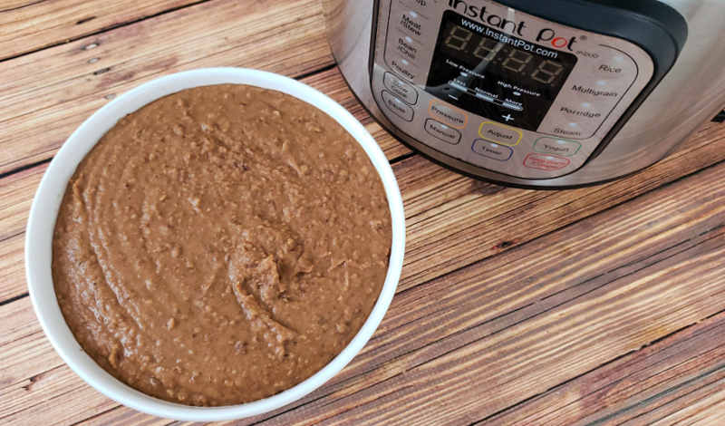 Refried beans in white ceramic bowl and Instant Pot on wood table.