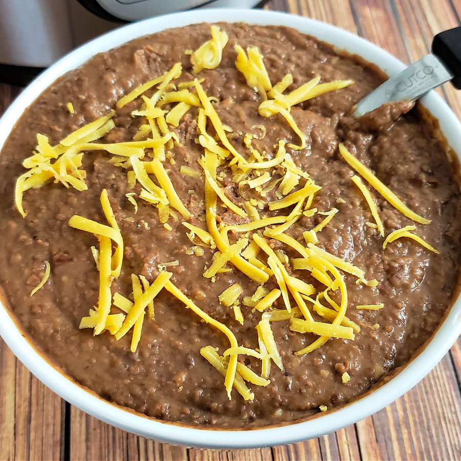 Close up of Instant Pot Refried Beans with shredded cheddar cheese in a white bowl with serving spoon.