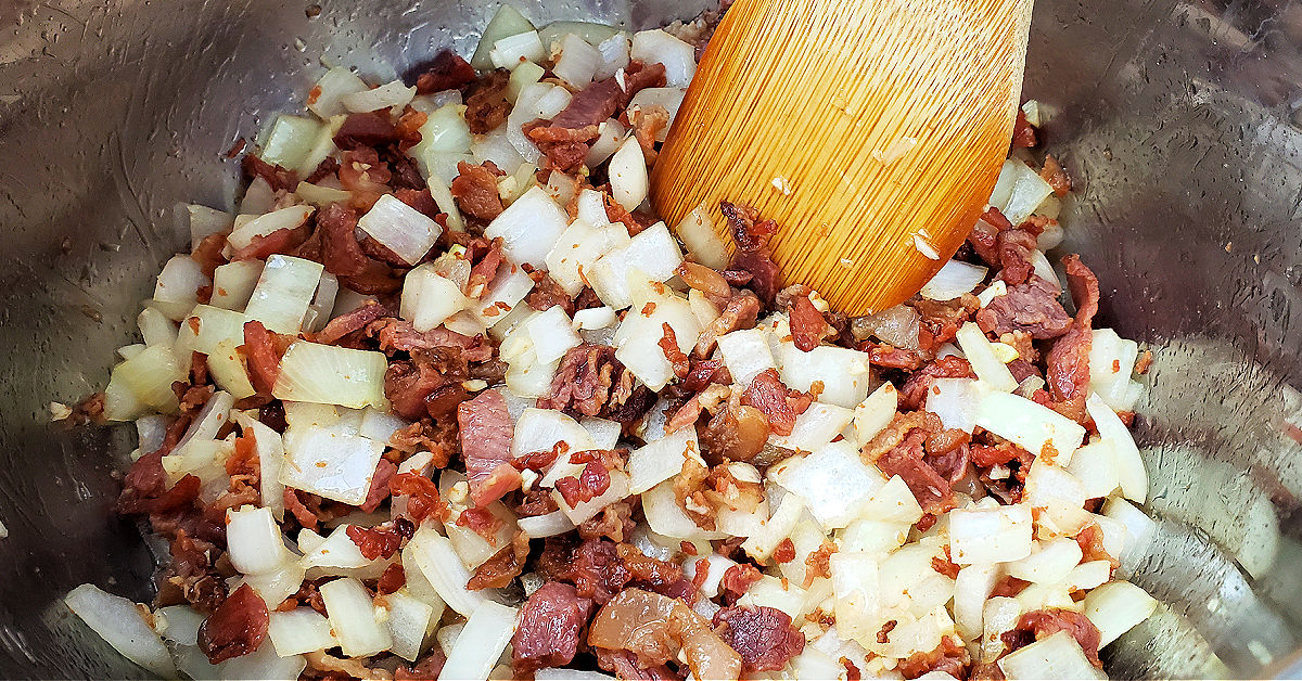 Onion and garlic being sauteed with cooked bacon in Instant Pot, being stirred with bamboo spatula.
