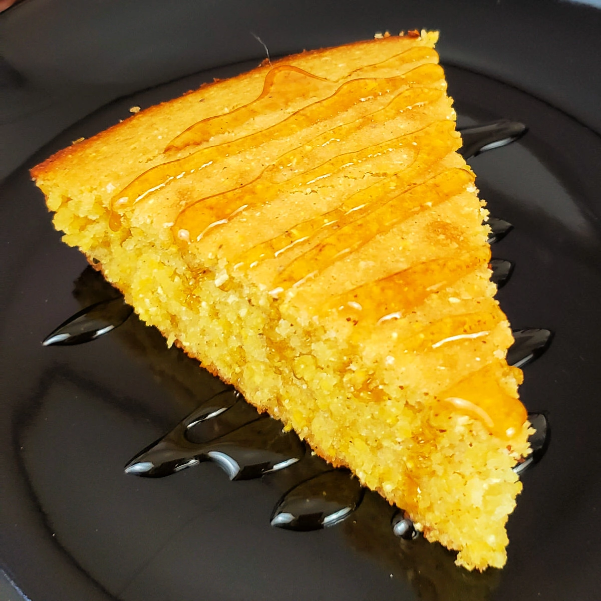 Slice of gluten free cornbread drizzled with honey on a black dessert plate.