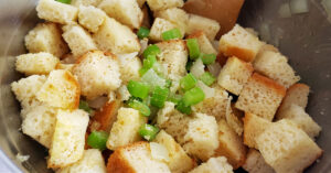 Celery, onion and fresh herbs added to bread cubes in bowl.