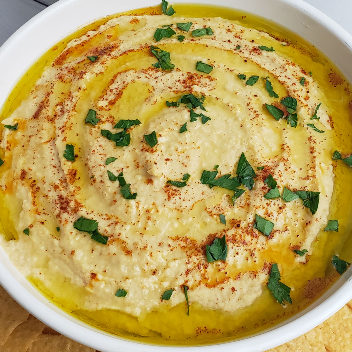 Delicious hummus garnished with extra virgin olive oil, fresh parsley and paprika, served in shallow white serving bowl with crackers.