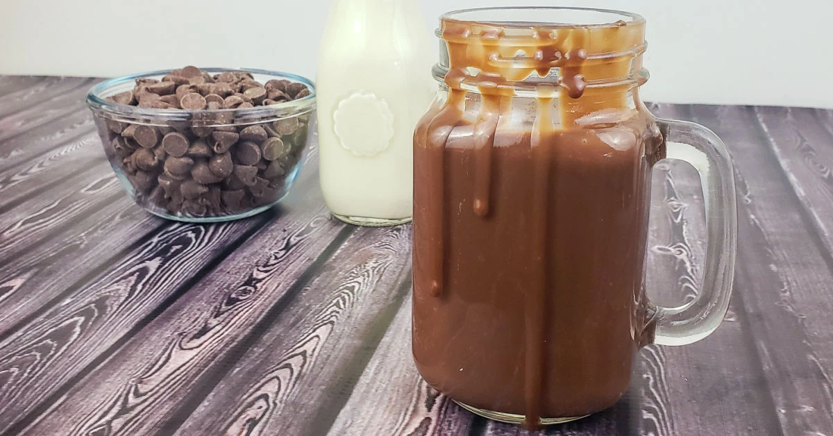 Jar of homemade chocolate sauce with cream and chocolate chips on table.