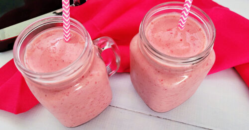 Two Strawberry Banana Smoothies served in pint size mason jars with handles and pink and white chevron paper straws next to a red napkin on a white table.