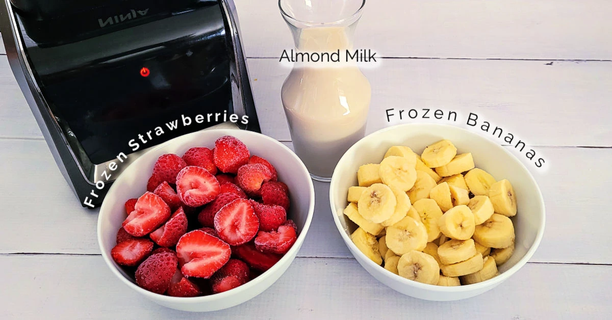 Ingredients for Strawberry Banana Smoothies, including 3 cups of frozen strawberries, 3 sliced frozen bananas and a pitcher of homemade almond milk next to a blender on a white table.