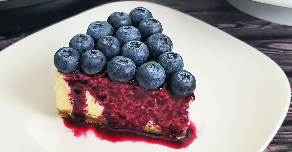 Gluten free cheesecake drizzled with blueberry syrup and topped with fresh blueberries on a white dessert plate.